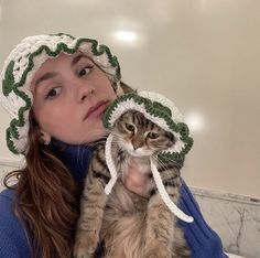 a woman is holding a cat wearing a crochet hat and looking at the camera