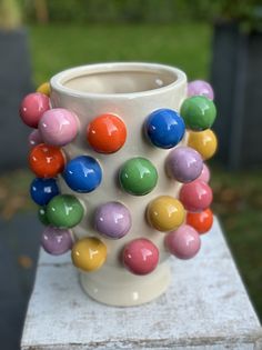 a white vase filled with lots of colorful balls on top of a wooden table next to grass