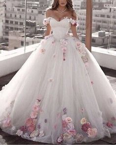 a woman in a white wedding dress with flowers on the skirt is standing near a window