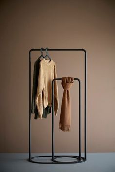 two sweaters hanging on clothes racks in front of a brown wall with a tan scarf