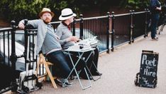 two men sitting at a table on the sidewalk with their arms around each other and one man wearing a hat