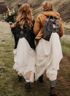 two women walking in the grass with backpacks on their back and flowers in their hands