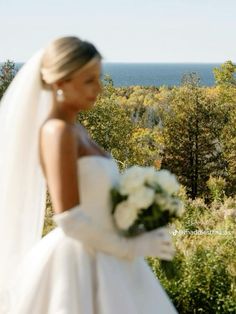 a woman in a white wedding dress holding a bouquet and looking out over the ocean