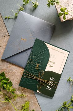 a green and gold wedding card on top of an envelope with greenery around it