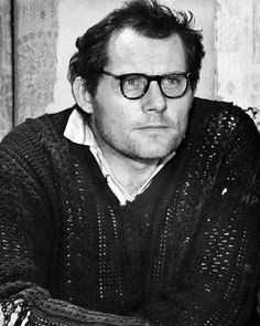 black and white photograph of a man wearing glasses sitting in front of a wooden wall