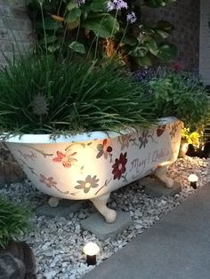 a bath tub filled with lots of plants next to some rocks and flowers on the ground