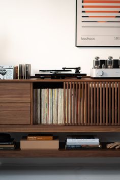 A modern wooden media console holding a record player, albums, and a stereo amplifier on top. The shelves below house vinyl records, books, and a small wooden figurine hand. On the wall above is a framed art print with geometric patterns. Record Player Tv Stand, Vinyl Player Setup, Vinyl Record Room Decor, Record Room Decor, Vinyl Setup, Modern Record Console, Record Player Setup, Record Player Aesthetic, Vinyl Cabinet