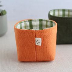 an orange and green fabric container with a white button on the side sitting on a table next to a potted plant