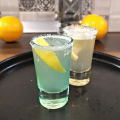 two drinks sitting on top of a black tray next to lemons and an orange