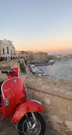 a red scooter parked on the side of a wall near water and boats