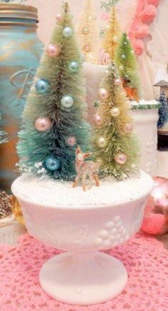 three small christmas trees in a white bowl on a pink doily tablecloth with other ornaments