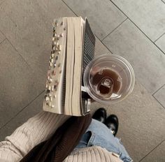 a person is holding an open book and a plastic cup with liquid in it while sitting on the floor