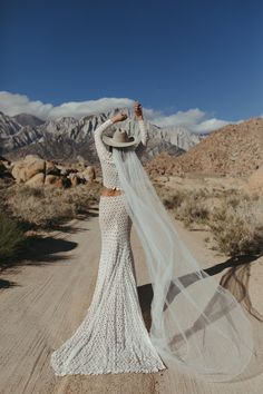 a woman in a white wedding dress and veil standing on a dirt road with mountains in the background