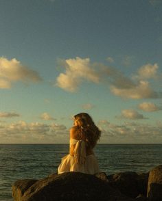 a woman sitting on top of a rock next to the ocean under a cloudy blue sky