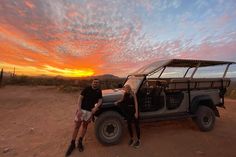 a man and woman standing next to a parked jeep in the desert at sunset or dawn