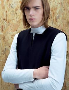 a young man with long hair wearing a black vest and white shirt, standing in front of a wood paneled wall