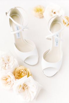 a pair of white high heeled shoes next to flowers on a white tablecloth