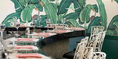 a long table with chairs and place settings in front of a wall painted with tropical leaves