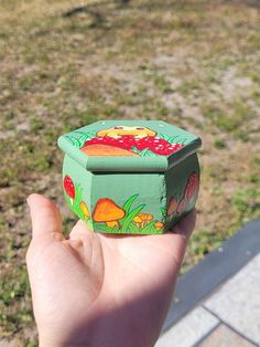 a hand holding a small green box with mushrooms painted on it's sides and grass in the background