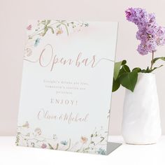 a white vase with purple flowers in it next to an open bar sign on a table