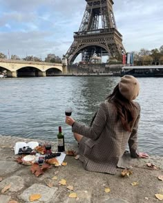 a woman sitting on the ground next to a body of water with a bottle of wine