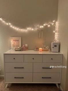 a white dresser topped with lots of drawers next to a wall covered in fairy lights