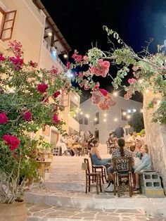 people are sitting at tables outside on the steps in front of some flowers and buildings