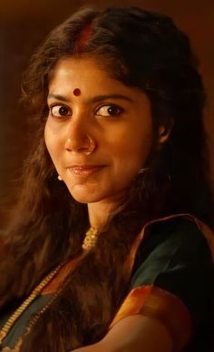 a woman with long dark hair wearing a green sari and gold necklace looking at the camera