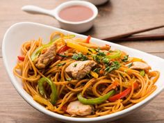 a white bowl filled with noodles and meat on top of a wooden table next to dipping sauce