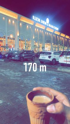 a person holding up a cup in front of a building with cars parked outside it