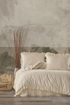 a white bed sitting in a bedroom next to a wooden trunk and vase with dry grass