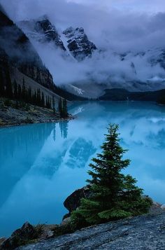 a blue lake surrounded by mountains with trees in the foreground and clouds in the background