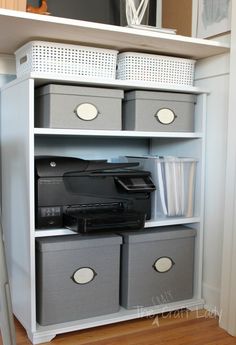 a white bookcase filled with gray bins next to a printer on top of a wooden floor