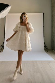 a woman standing in front of a white backdrop wearing a dress and holding a cell phone to her ear