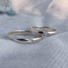 two white gold wedding bands with diamonds on them sitting on top of a cloth covered blanket