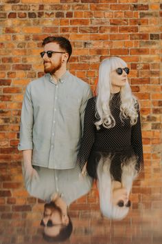 a man and woman standing next to each other in front of a brick wall wearing sunglasses