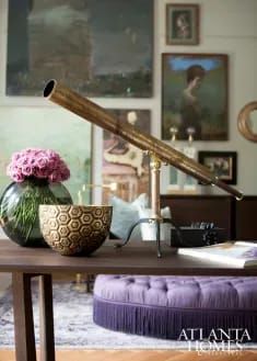 a table with a purple ottoman and flowers in a vase next to an old telescope