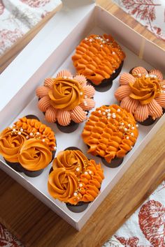 six cupcakes with orange frosting in a white box on a wooden table