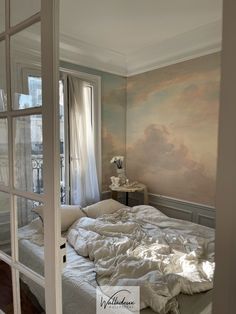 an unmade bed sitting next to a window in a room with a painting on the wall