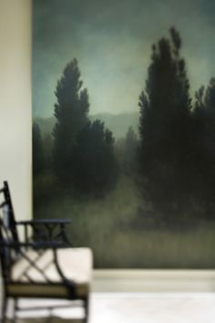 a chair sitting in front of a painting with trees on the wall behind it,