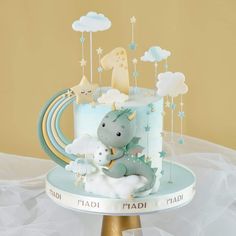 a blue cake decorated with an elephant and baby's name on the top is surrounded by clouds