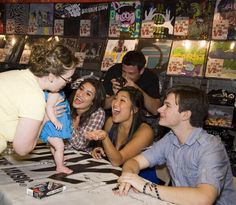 a group of people sitting around a table with a baby in the middle and an adult standing next to them