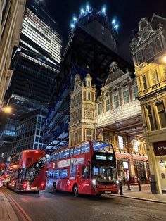 two red double decker buses driving down the street in front of tall buildings at night