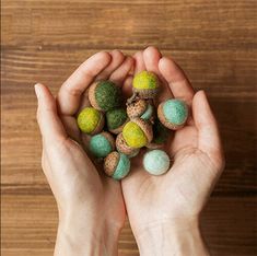 two hands are holding small balls of colored rocks on a wooden surface, one is green and the other is blue