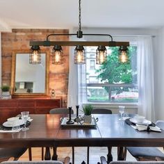 a dining room table and chairs with lights hanging from the ceiling over it, in front of a brick wall