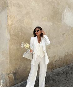 a woman standing next to a wall wearing white pants and a blazer with flowers in her hand