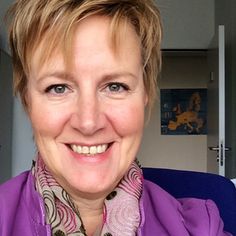 a woman with short hair wearing a purple shirt and scarf smiles at the camera while sitting in an airplane