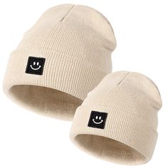 two white knit hats with smiley faces on the front and one has a black patch