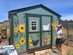 a woman painting the side of a shed with sunflowers and chickens on it