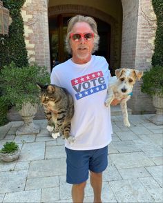 a man holding two cats and a dog in front of a building with an american flag t - shirt